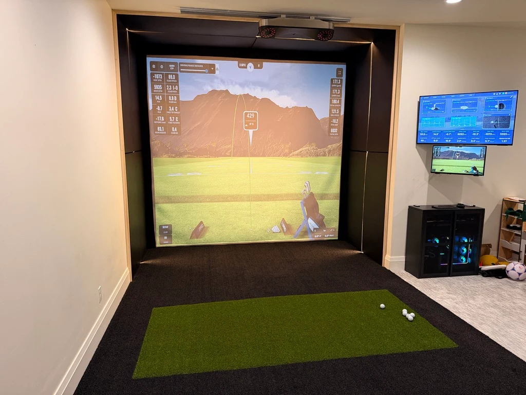 Custom home golf simulator with a padded enclosure, overhead launch monitor, and dual wall-mounted displays